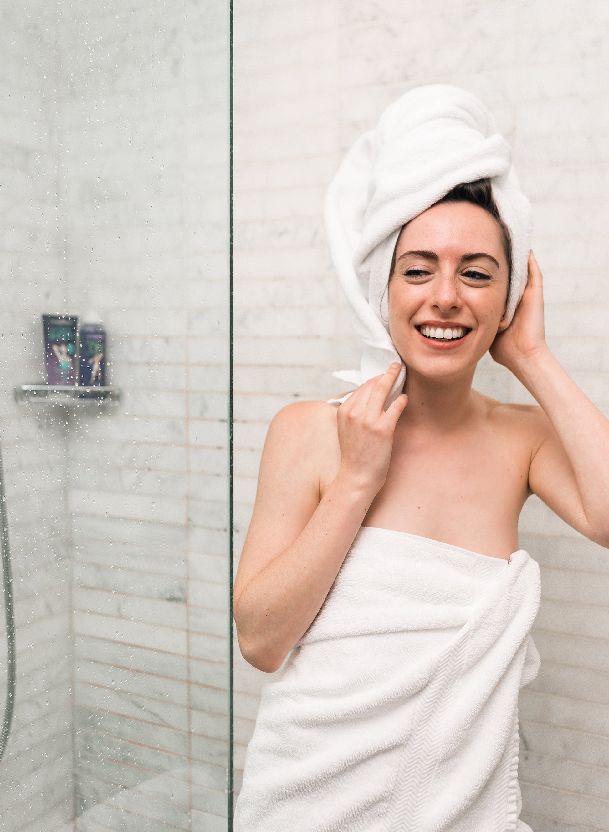 A smiling woman stepping out of a shower, wrapped in a towel with another towel around her hair.