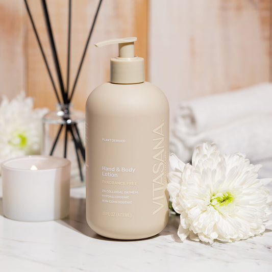 16 fl oz pump bottle of Vitasana Fragrance-Free Hand & Body Lotion, sitting on a white countertop with a white flower, rolled up towels, a candle, and incense sticks. Bottle features the Vitasana logo on a pale tan background.